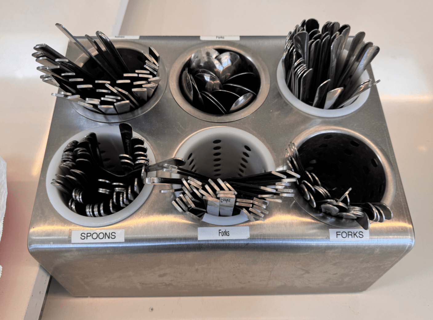 A stainless steel dining hall caddy with six identical round bins. Labels say spoons and forks, but the utensils inside do not match.