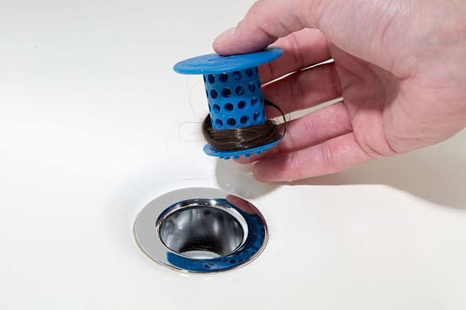 A hand holding a blue drain catcher wrapped in hair, above an open shower drain